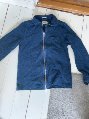 Blå overshirt med dragkedja - Säljer en blå overshirt från East West med dragkedja framtill och klassisk krage. Overshirten har en bröstficka och är tillverkad i ett mjukt, jeansliknande material. Perfekt att slänga på över en t-shirt för en avslappnad look.