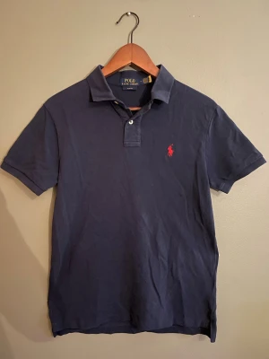 Marinblå pikétröja från Polo Ralph Lauren - Klassisk marinblå pikétröja från Polo Ralph Lauren med korta ärmar och röd broderad logga på bröstet. Tröjan har krage och två knappar vid halsen. Perfekt till jeans eller shorts för en clean och stilren look.