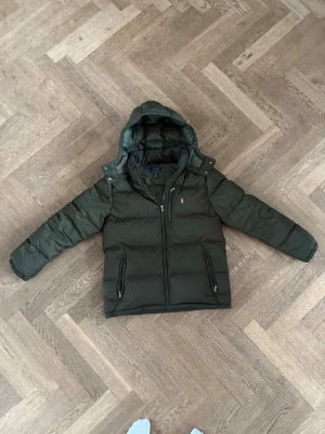 Grön dunjacka från Polo Ralph Lauren - Snygg mörkgrön dunjacka från Polo Ralph Lauren med huva och dragkedja framtill. Jackan har två dragkedjefickor, diskret logga på bröstet och quiltad design. Perfekt för kalla vinterdagar och riktigt skön att bära.