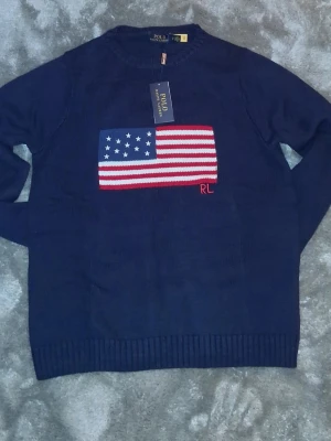 Mörkblå stickad tröja Polo Ralph Lauren - 2 styckna Snygga mörkblå stickad tröja från Polo Ralph Lauren med amerikansk flagga i rött, vitt och blått på bröstet samt RL-broderi. Tröjan har rund halsringning och ribbstickade muddar. Perfekt för dig som gillar klassisk preppy stil.