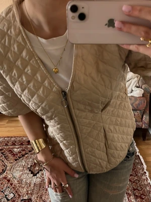 Beige quiltad cardiganjacka - Säljer en beige quiltad cardiganjacka med dragkedja framtill och korta ärmar. Jackan har en avslappnad passform och är tillverkad i ett lätt vadderat material som ger en cool look. Perfekt att slänga över en t-shirt för en trendig vibe.