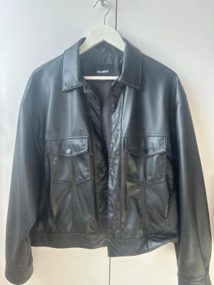 Svart skinnjacka från Pull&Bear - Snygg svart skinnjacka från Pull&Bear med klassisk krage och två stora bröstfickor med lock och knapp. Rak passform och stilrena detaljer gör den enkel att matcha till olika outfits. Perfekt för dig som gillar en tidlös och cool look. Jackan är S oversized men passar som M också.