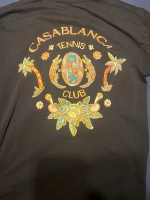 Svart Casablanca Tennis Club t-shirt - Snygg svart t-shirt från Casablanca med färgglad 'Tennis Club'-print på både bröstet och ryggen. Trycket har detaljer med palmer, tennisbollar och blommor i grönt, gult och orange. Klassisk rund hals och korta ärmar. Perfekt för dig som gillar streetwear med sportig vibe.