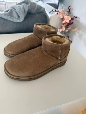 Bruna fårskinnstofflor från UGG - Mysiga bruna tofflor från UGG i klassisk modell med mjukt fårskinnsfoder och mocka på utsidan. De har en låg ankelhöjd och bred passform, perfekta för att hålla fötterna varma hemma. UGG-logga bak på hälen och robust sula för extra komfort.