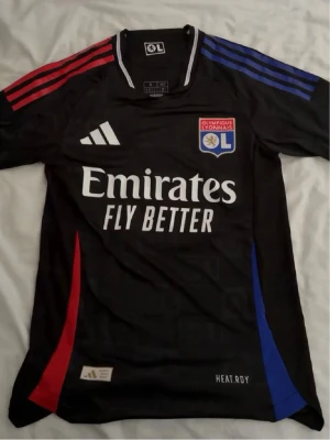 Olympique Lyonnais svart matchtröja - Snygg svart matchtröja från Olympique Lyonnais med Adidas-logga och klubbmärke på bröstet. Tröjan har röda och blå detaljer på axlar och sidor samt vit text med Emirates Fly Better på framsidan. Tillverkad i funktionsmaterial med HEAT.RDY-teknologi.