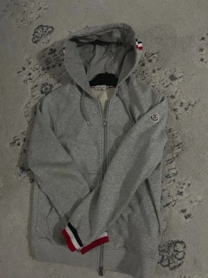 Grå zip hoodie från Moncler - Snygg grå hoodie från Moncler med dragkedja framtill, huva med dragsko och fickor på sidorna. Ärmslutet har Monclers signaturfärger i rött, vitt och blått. Moncler-logga på ärmen och diskret flaggdetalj på huvan. Perfekt för en avslappnad streetwear-look.