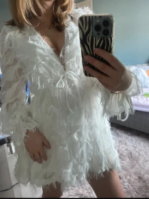 Studentklänning  - Supersöt vit kortklänning med fjäderliknande detaljer och v-ringning. Klänningen har långa ärmar och snörning framtill som ger en extra touch. Perfekt för sommaren eller studenten! Prislapp kvar🥰