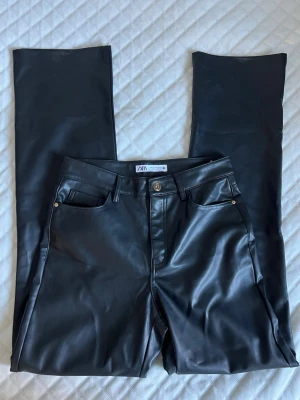 Svarta coated jeans från Zara - Snygga svarta coated jeans från Zara med raka ben. Jeansen har en glansig finish som ger en cool läderlook och detaljer i guld vid fickorna. Använda fåtal gånger, små slitningar nere vid foten, knappt synligt. 