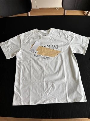 Vit Maison Margiela t-shirt - Vit t-shirt från Maison Margiela med tryckt logga och siffror på bröstet. Klassisk passform med rund hals och korta ärmar. Tillverkad i mjuk bomull för en skön känsla. Perfekt för dig som gillar minimalistisk och stilren design.