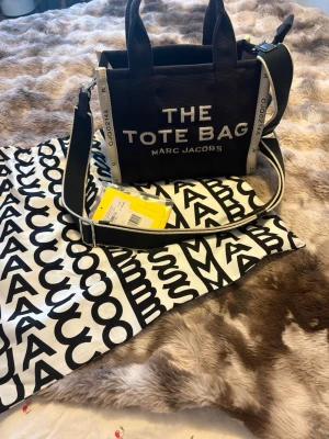  Tote Bag från Marc Jacobs - Säljer en svart shoppingväska från Marc Jacobs med texten 'THE TOTE BAG' framtill. Väskan har vita detaljer, bred axelrem och är tillverkad i canvas. Medföljer dustbag med svartvitt mönster och logga. Köpt på miinto har digitalt kvitto 