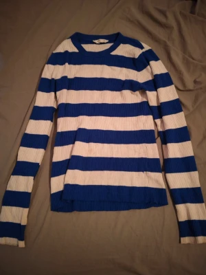 Blåvit randig stickad tröja Gina Tricot - Säljer en ribbstickad tröja från Gina Tricot med breda blå och vita ränder. Tröjan har rund halsringning och långa ärmar. Perfekt för dig som gillar klassiska marininspirerade plagg med en modern twist.