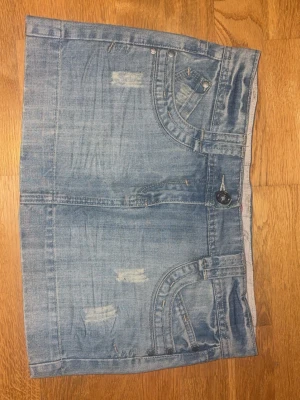 Blå jeanskjol med slitningar - Säljer en blå jeanskjol i kort modell med slitna detaljer och klassiska fickor både fram och bak. Kjolen har knapp och dragkedja framtill samt tydliga sömmar för en cool denimlook. Perfekt för dig som gillar en avslappnad och trendig stil.