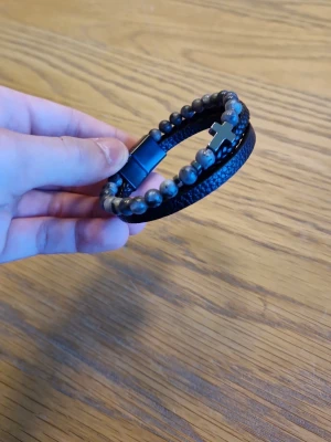 Svart armband med pärlor och kors - Stilrent svart armband med mattsvarta pärlor och ett metallkors som detalj. Armbandet har även en flätad läderrem och magnetlås. Perfekt accessoar för dig som gillar en edgy och modern look.