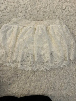 Vit spetskjol med volang - Söt vit kortkjol i spets med volangkant. Kjolen har ett romantiskt blommigt spetsmönster och resår i midjan för skön passform. Perfekt att styla med en enkel topp för en trendig look.