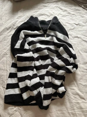 Svartvit randig stickad tröja - Stickad tröja med breda svarta och vita ränder, halvzip i halsen och ribbstickade muddar. Oversized passform och mjukt material, perfekt för lager på lager. Klassisk och trendig design som passar till jeans eller kjol.