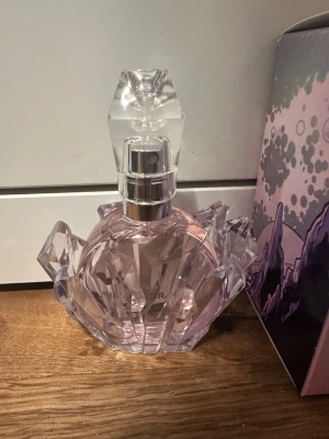 Ariana Grande parfym i kristallflaska - Unik parfym från Ariana Grande i en rund, ljusrosa flaska med kristalliknande detaljer och silvrig spraydel. Flaskan har en cool, futuristisk design och kommer i en snygg förpackning med tecknad illustration. Aldrig använt, orginal pris är 595kr.