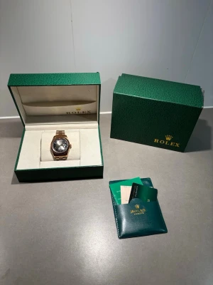  guldarmbandsur med svart urtavla - Lyxig klocka med guldfärgat armband och svart urtavla. Kommer i original grön box med certifikat och tillbehör. Klassisk design med datumfönster och räfflad bezel, perfekt för dig som vill ha något exklusivt på handleden.