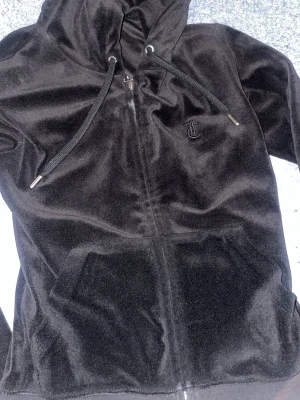 Svart velour hoodie med dragkedja - Säljer en svart hoodie från Juicy Couture i mjuk velour. Hoodien har dragkedja framtill, fickor och justerbar huva med snören. Klassisk JC-logga broderad på bröstet. Perfekt för en chill och trendig look. Juicy couture loggan på koftan är lite sönder men funkar fortfarande 