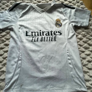 Real Madrid Mbappé matchtröja vit - Säljer en vit Real Madrid matchtröja från Adidas med svarta detaljer och klubbmärke på bröstet. Tryck med 'Emirates Fly Better' på framsidan och 'Mbappé 9' på ryggen. Perfekt för fotbollsfans som vill sticka ut på planen eller läktaren.