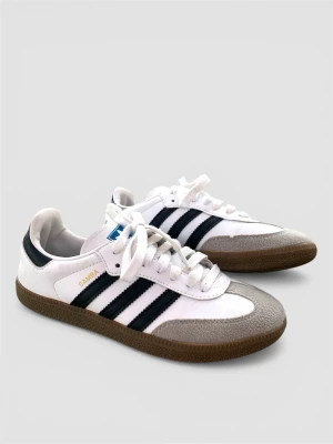 Adidas Samba vita och svarta sneakers - Klassiska Adidas Samba sneakers i vitt läder med svarta detaljer och de ikoniska tre ränderna på sidan. Skon har brun gummisula och grå mockadetaljer vid tån. Guldtexten 'SAMBA' syns på sidan. Perfekt retrostil för dig som gillar streetwear.