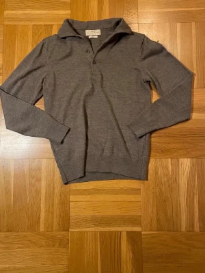 Grå stickad tröja från The Tailoring Club - Säljer en stilren grå stickad tröja från The Tailoring Club. Tröjan har krage och två knappar vid halsen, vilket ger en klassisk look. Materialet är mjukt och tunt, perfekt för lager-på-lager. Långa ärmar och ribbade muddar vid ärmslut och nederkant.