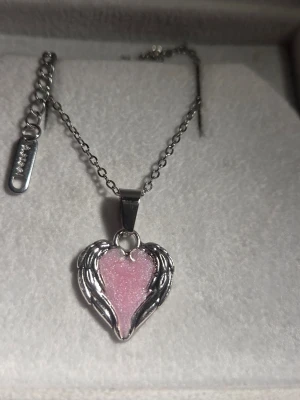 Halsband med rosa självlysande hjärthänge och vingar - Snyggt halsband i silverfärgad metall med ett hjärtformat hänge i rosa glitter och detaljerade vingar runt hjärtat som lyser blått i mörker efter att ha varit i dagsljus eller solljus. Kedjan är tunn i rostfritt stål och har justerbar längd. Perfekt accessoar för dig som gillar unika smycken med lite extra attityd. Både kedjan och hänget tål vatten och missfärgas inte.