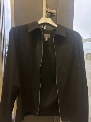 Svart overshirt från H&M med dragkedja - Stilren svart overshirt från H&M med klassisk krage, dragkedja framtill och bröstficka. Materialet är slätt och lätt, perfekt för lager-på-lager. Långa ärmar med knappar vid manschetterna ger en snygg och enkel look.