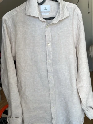 Beige linneskjorta från Bläck - Snygg beige skjorta från Bläck i linnematerial med klassisk krage och knappar framtill. Skjortan har lång ärm och en skräddarsydd passform som ger en clean och fräsch look. Perfekt för dig som gillar stilrena plagg med en avslappnad vibe.