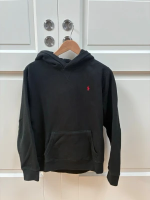 Svart hoodie från Polo Ralph Lauren - Klassisk svart hoodie från Polo Ralph Lauren med röd broderad logga på bröstet. Hoodien har huva, känguruficka framtill och ribbade muddar vid ärmar och nederkant. Tillverkad i mjukt bomullsmaterial som är skönt att bära.