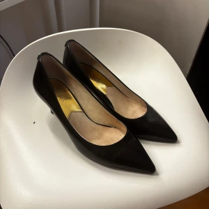 Svarta lackpumps med gulddetalj - Snygga svarta pumps i lack med spetsig tå och låg klack. Guldfärgad detalj bak på hälen och beige innersula. Klassisk och elegant modell som passar till många olika outfits.