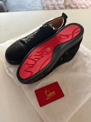 Svarta Louboutin sneakers med röd sula - Snygga svarta sneakers från Christian Louboutin med ikonisk röd sula och diskret logga på sidan. Skorna har lågt skaft, svarta skosnören och är tillverkade i mocka och läder. Perfekta för dig som vill sticka ut med exklusiv stil.
