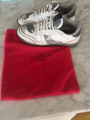  Snygga skor  - Snygga sneakers från  i vitt, grått och svart med camomönster. Skorna har lågt skaft, klassisk snörning och detaljer i mocka och läder. Sulan är svart och har en unik nitar-detalj vid hälen. Perfekta för dig som gillar exklusiva streetwear-vibbar.sulan lite slitig och några märken på skon 