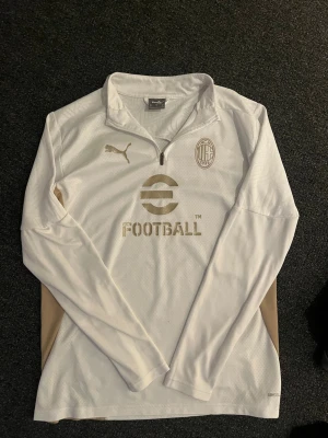 Vit AC Milan träningströja Puma - Snygg vit långärmad träningströja från Puma med AC Milan-logga och guldiga detaljer. Tröjan har halv dragkedja framtill, guldiga inslag på sidorna och är tillverkad i ett lätt funktionsmaterial. Perfekt för fotbollsträning eller sportiga dagar.