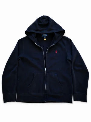 Svart hoodie från Polo Ralph Lauren - Svart hoodie med dragkedja från Polo Ralph Lauren. Klassisk design med huva, kängurufickor och det ikoniska röda Polo-logotrycket på bröstet. Tillverkad i mjukt bomullsmaterial som är skönt att ha på sig. Perfekt för en avslappnad och stilren look.