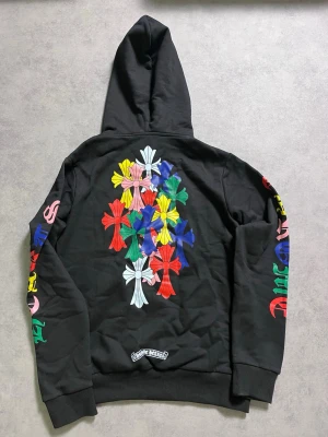 Chrome hearrs - Svart hoodie från Chrome Hearts med broderade färgglada kors på ryggen och mindre kors på bröstet. Ärmarna har tryck i olika färger och huva med dragsko. Perfekt för dig som gillar streetwear och unika detaljer. 