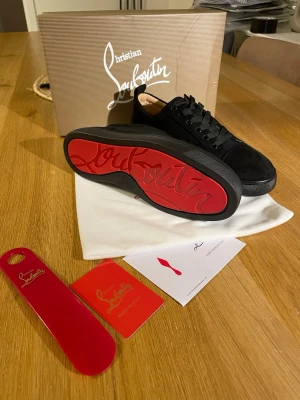 Svarta Louboutin sneakers med röd sula - Stilrena svarta sneakers från Christian Louboutin i mocka med klassisk röd sula och svarta skosnören. Låg modell med diskret logga på sulan och snygga detaljer. Perfekta för dig som vill ha något exklusivt och trendigt.