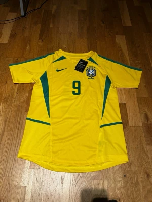Brazil 2002 Ronaldo #9 - Gul Brasilien fotbollströja från Nike med gröna detaljer, nummer 9 och Ronaldo på ryggen. Klassisk design med korta ärmar och rund hals. Perfekt för dig som älskar fotboll och vill hylla en ikonisk spelare.