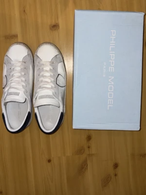 Vita Philippe Model  - Snygga vita sneakers från Philippe Model med grå mockadetaljer och mörkblå häl. Klassisk låg modell med vita skosnören och ikonisk sköldlogga på sidan. Skorna har en stilren design och passar perfekt till streetwear och casual outfits. Skorna är använda men har inga skador, bara lite smutsiga. Tvättar man dem är de som nya! Väldigt eftertraktade skor. Priset är ej hugget i sten. Var inte rädda att ställa frågor!😁