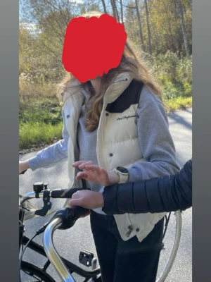 Vit dunväst från Peak Performance - Snygg vit dunväst från Peak Performance med bruna axelpartier och broderad logga på bröstet. Västen har hög krage, dragkedja och tryckknappar framtill samt två dragkedjefickor. Perfekt för lager på lager och riktigt skön att bära utomhus. I bra skick men syns att den är använd. Två mindre fläckar på framsidan samt lite slitage på ”lädret” (se bilder).