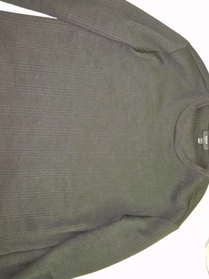 Svart stickad tröja från SMOG - En stilren svart stickad tröja från SMOG med rund halsringning och diskret ribbad struktur. Tröjan har lång ärm och en klassisk passform som funkar till det mesta. Perfekt för dig som gillar enkel och clean stil.