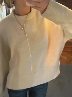 Beige oversized stickad tröja - Mysig beige stickad tröja med oversized passform och breda ribbade muddar vid ärmslut och nederkant. Tröjan har en enkel design med en dekorativ söm framtill och är perfekt för lager-på-lager-stil. Materialet ser ut att vara mjukt och värmande.