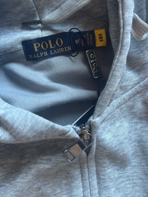 Grå hoodie från Polo Ralph Lauren - Snygg grå hoodie från Polo Ralph Lauren med dragkedja framtill och klassisk svart logga broderad på bröstet. Hoodien har huva med dragsko och är tillverkad i mjukt bomullsmaterial. Perfekt för en chill och stilren look.