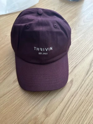 Vinröd keps från Thrivin - En stilren vinröd keps från Thrivin med broderad vit text framtill. Klassisk böjd skärm och justerbart spänne bak. Tillverkad i bomullstyg som känns både mjukt och slitstarkt. Perfekt accessoar för en avslappnad streetwear-look. Oanvänd!