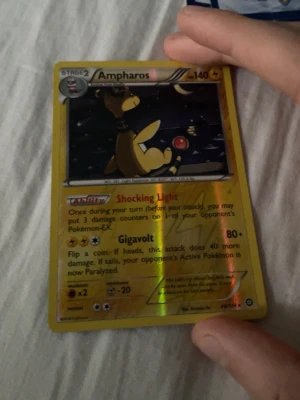 Ampharos Pokémonkort holografiskt - Säljer ett holografiskt Ampharos Pokémonkort med gul ram och svart bakgrund. Kortet har 140 HP och attacker som Shocking Light och Gigavolt. Perfekt för samlare eller dig som vill boosta din lek med en el-Pokémon.