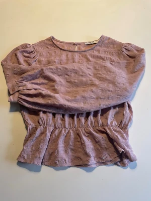 Ljuslila blus med puffärm från And - Söt ljuslila blus från And med puffiga långärmar och rund halsringning. Blusen har ett strukturerat tyg och smockad midja som ger en fin siluett. Perfekt att styla med jeans eller kjol för en trendig look.
