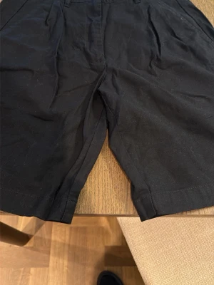Svarta shorts från 157 - Säljer ett par svarta shorts från lager 157 med klassisk knappstängning. Shortsen har en rak passform och är tillverkade i bomull, vilket gör dem både bekväma och stilrena. Perfekta för varma dagar och enkla att matcha med olika outfits. Nytt pris 150kr