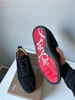 Christian Louboutin svarta spikes sneakers  - Svarta sneakers i mocka från Christian Louboutin med svarta nitar på tån och klassisk röd sula med logga. Skorna har svarta skosnören och en låg silhuett. Perfekta för dig som vill sticka ut med en edgy och lyxig stil. Inget skohorn följer med. 