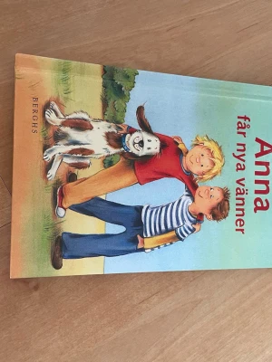 Anna får nya vänner bok - En färgglad barnbok med titeln 'Anna får nya vänner' från Berghs. Omslaget visar två barn och en hund i en lekfull scen. Boken har hårda pärmar och är illustrerad med stora bilder och tydlig text, perfekt för yngre läsare.