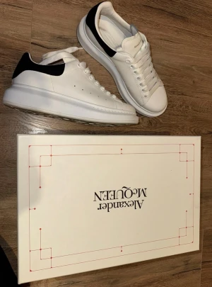 Vita Alexander McQueen sneakers - Stilrena vita sneakers från Alexander McQueen med svart hälparti och tjock sula. Klassisk låg modell med perforerade detaljer på sidan och vita skosnören. Tillverkade i läder och levereras med originalkartong och extra skosnören.