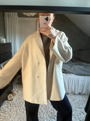 Oversized vit ullkappa - Stilren oversized kappa i vit ullmix med dubbelknäppning och breda slag. Jackan har en rak passform och släta knappar framtill. Perfekt för dig som gillar minimalistisk och trendig stil.